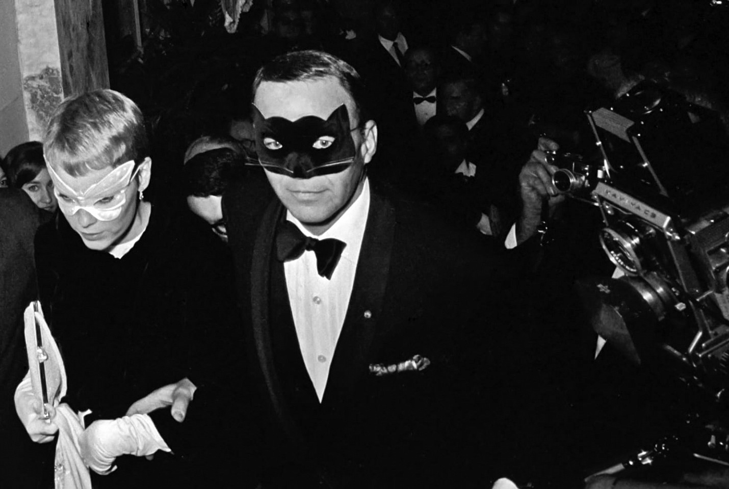 Harry Benson, Z Frank Sinatra and Mia Farrow, Truman Capote Ball; edition of 35, 1966
Photograph, 40 x 60 inches $22,500,  30 x 40 inches $20,000,  24 x 30 inches $18,000,  20 x 24 inches $15,000, 17 x 22 inches $12,000
Harry Benson’s photograph captures a moment of glamour and intrigue as Frank Sinatra and Mia Farrow make their entrance into Truman Capote’s legendary Black and White Ball at the Plaza Hotel in NYC in 1966. The atmosphere is electric with anticipation, as the couple, adorned in elegant masks, step into the spotlight.

Mia Farrow’s mask, a delicate white butterfly, contrasts beautifully with Frank Sinatra’s striking black cat mask complete with whiskers. Sinatra, with a smirk on his face, gazes directly at the camera, exuding confidence and charisma as he leads Farrow into the festivities. The couple’s connection is palpable, their clasped hands and Sinatra’s guiding gesture suggesting a sense of unity and mutual support.

In the background, a throng of photographers and onlookers capture the moment, underscoring the immense public interest and intrigue surrounding the event. Tuxedoed men mingle in the background, adding to the aura of sophistication and exclusivity that permeates the scene.

Benson’s photograph encapsulates the allure and mystique of Capote’s Black and White Ball, an event that transcended mere celebrity to become a cultural phenomenon. The party, attended by the elite of society, was the hottest ticket in town, with exclusivity so coveted that even those who were not invited would fabricate excuses rather than admit their absence.

The occasion was held in honor of Kay Graham, owner of the Washington Post, and the detectives who cracked the murder case depicted in Capote’s groundbreaking novel, “In Cold Blood.” For Capote, it was a moment of triumph, a pinnacle of his literary and social success.

Benson’s photograph serves as a vivid snapshot of a bygone era, capturing the essence of an iconic event that continues to captivate imaginations decades later. In the faces of Sinatra and Farrow, we glimpse the allure of fame and the magnetic pull of a night steeped in glamour and intrigue.
BENS00029